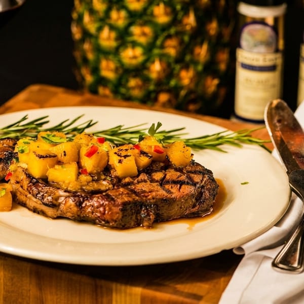 Grilled steak topped with pineapple salsa and rosemary, with a glass of wine and pineapple in the background.