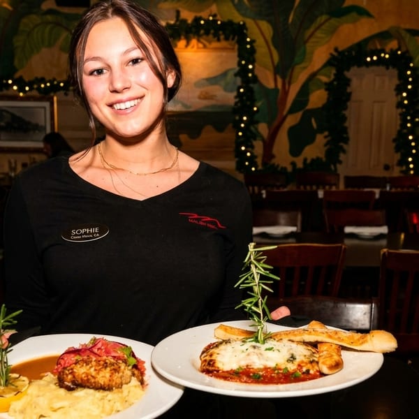 Smiling server Sophie holds two plates of Italian-American food in a dimly lit restaurant dining room.