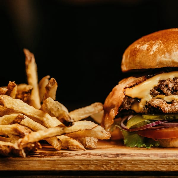 Cheeseburger with two patties, lettuce, tomato, onion, pickles, and a side of fries on a wooden board.