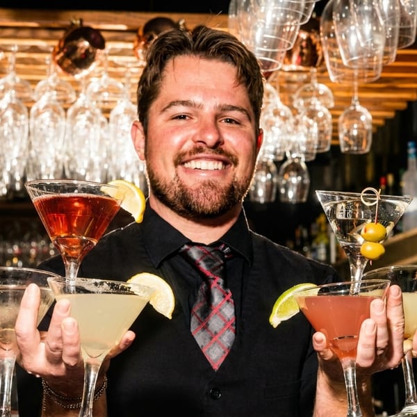 Smiling bartender holding five colorful cocktails in martini glasses at a bar.