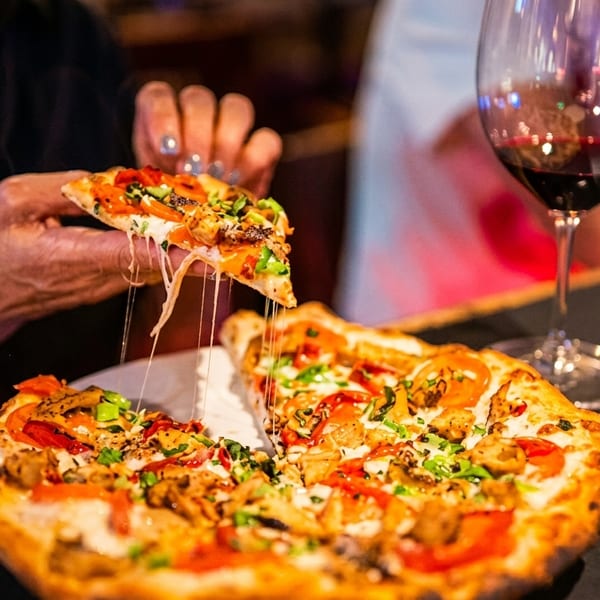 A hand pulls a slice of pizza with melted cheese strings, next to a glass of red wine.